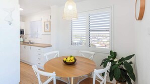 A dining area with a wooden table, white chairs, a bowl of fruit, a hanging light fixture, and a window with blinds. - Poppy's Place - Oceanstays (Yamba)