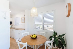 A dining area with a wooden table, white chairs, a bowl of fruit, a hanging light fixture, and a window with blinds.