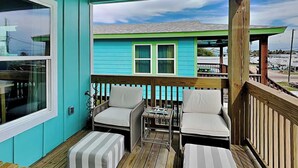 A covered outdoor deck with two cushioned chairs and a small table, wooden flooring, and a view of a blue house with green trim. - Marlin Manor by Silver Sands Vacation Rentals (Port Aransas)