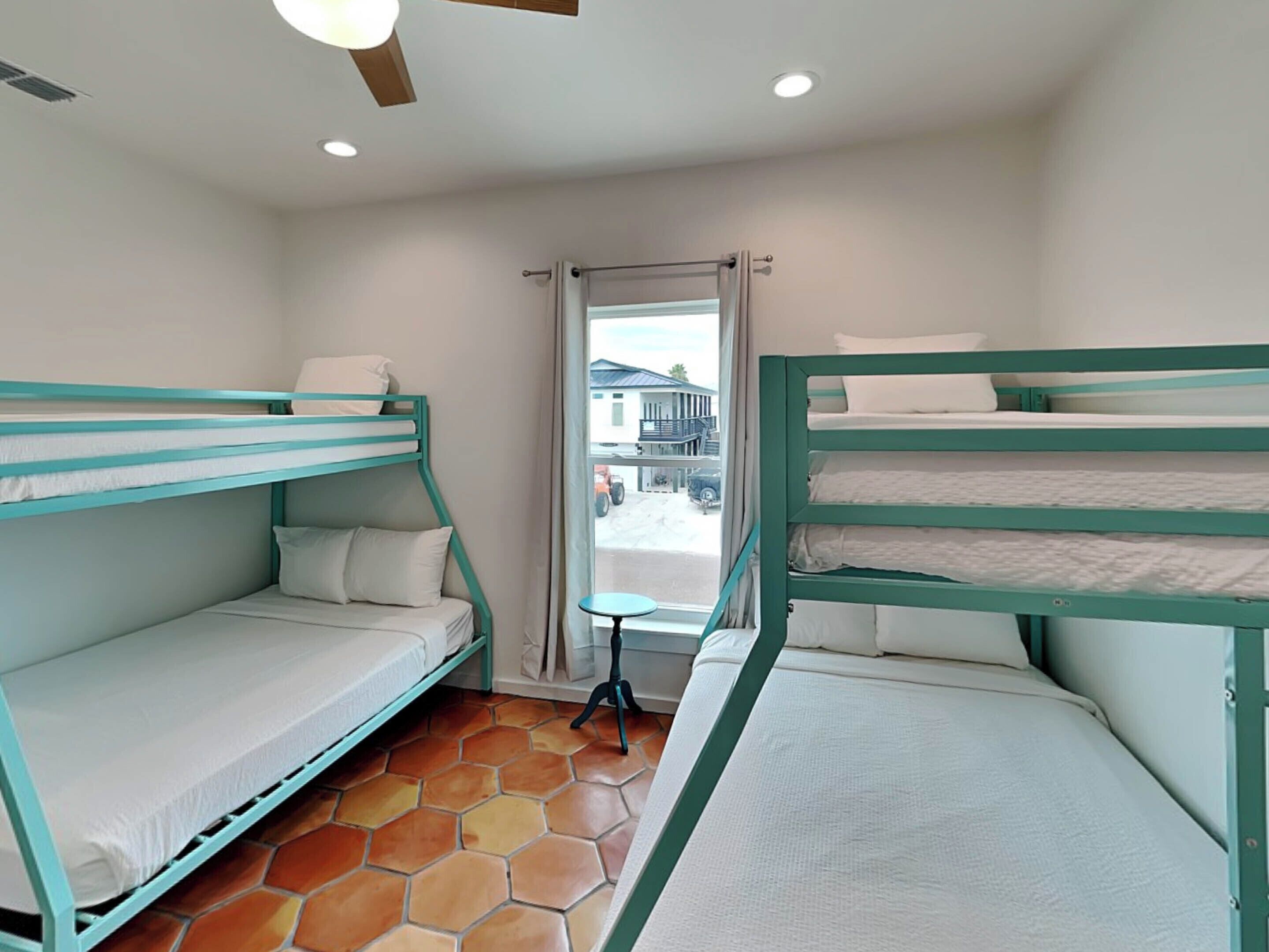 A dormitory room with bunk beds, a tiled floor, a window with a view of a building and vehicles outside, and a ceiling fan.