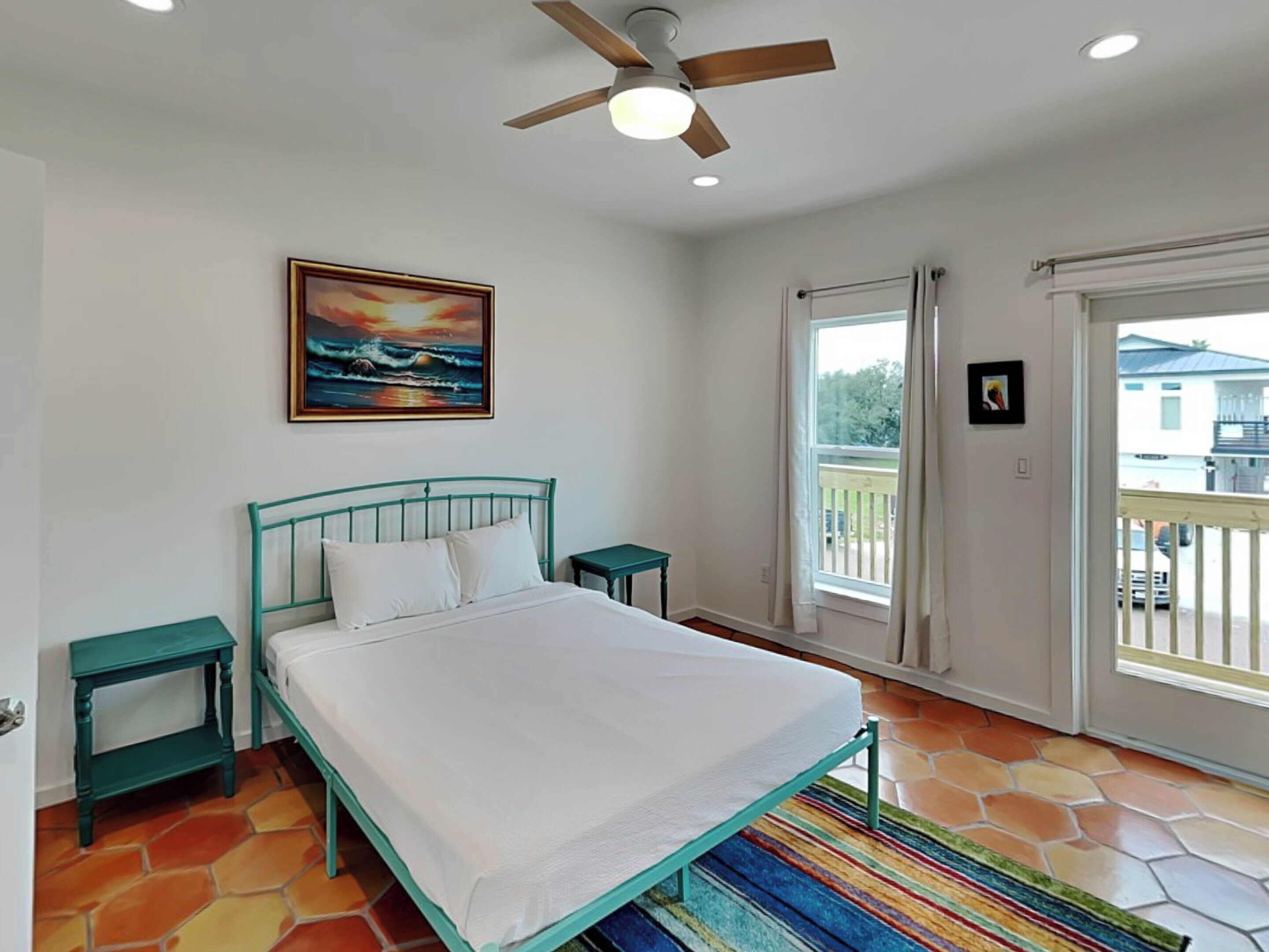 A bedroom with a bed, a ceiling fan, a painting, a window with curtains, and a view of the outside.