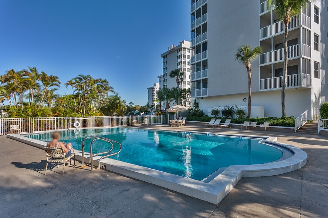 A swimming pool with a curved edge, surrounded by a white fence. There are lounge chairs and a palm tree near the pool.