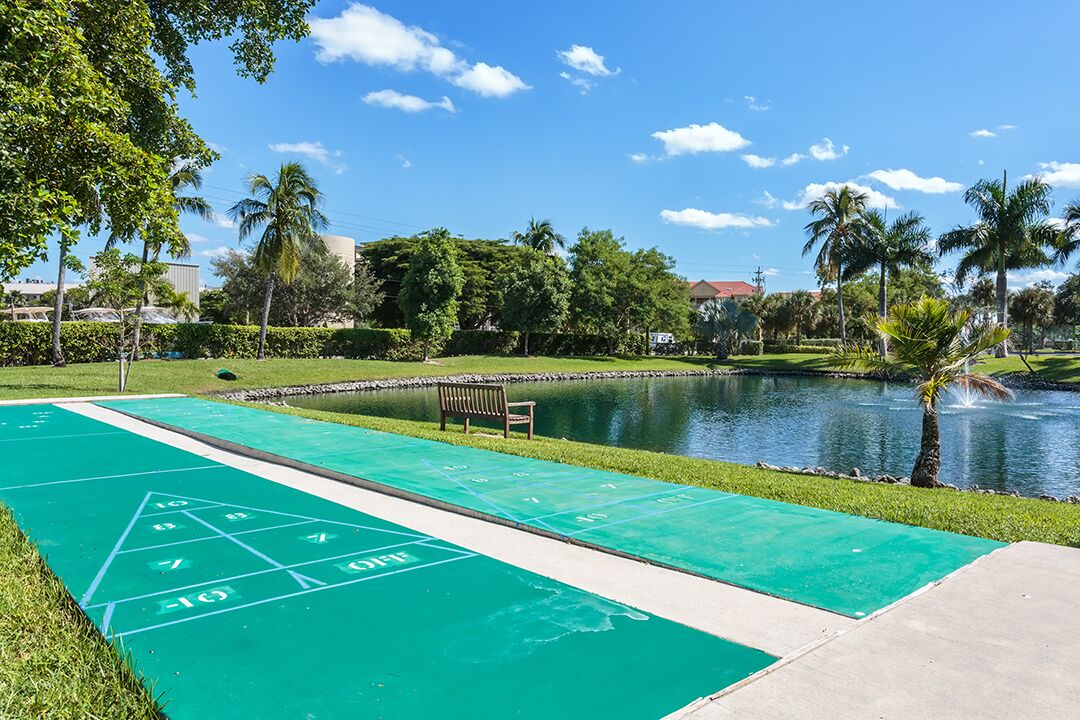 Un terrain de jeu de shuffleboard vert, situé à côté d’une étendue d’eau, avec des palmiers et un banc.