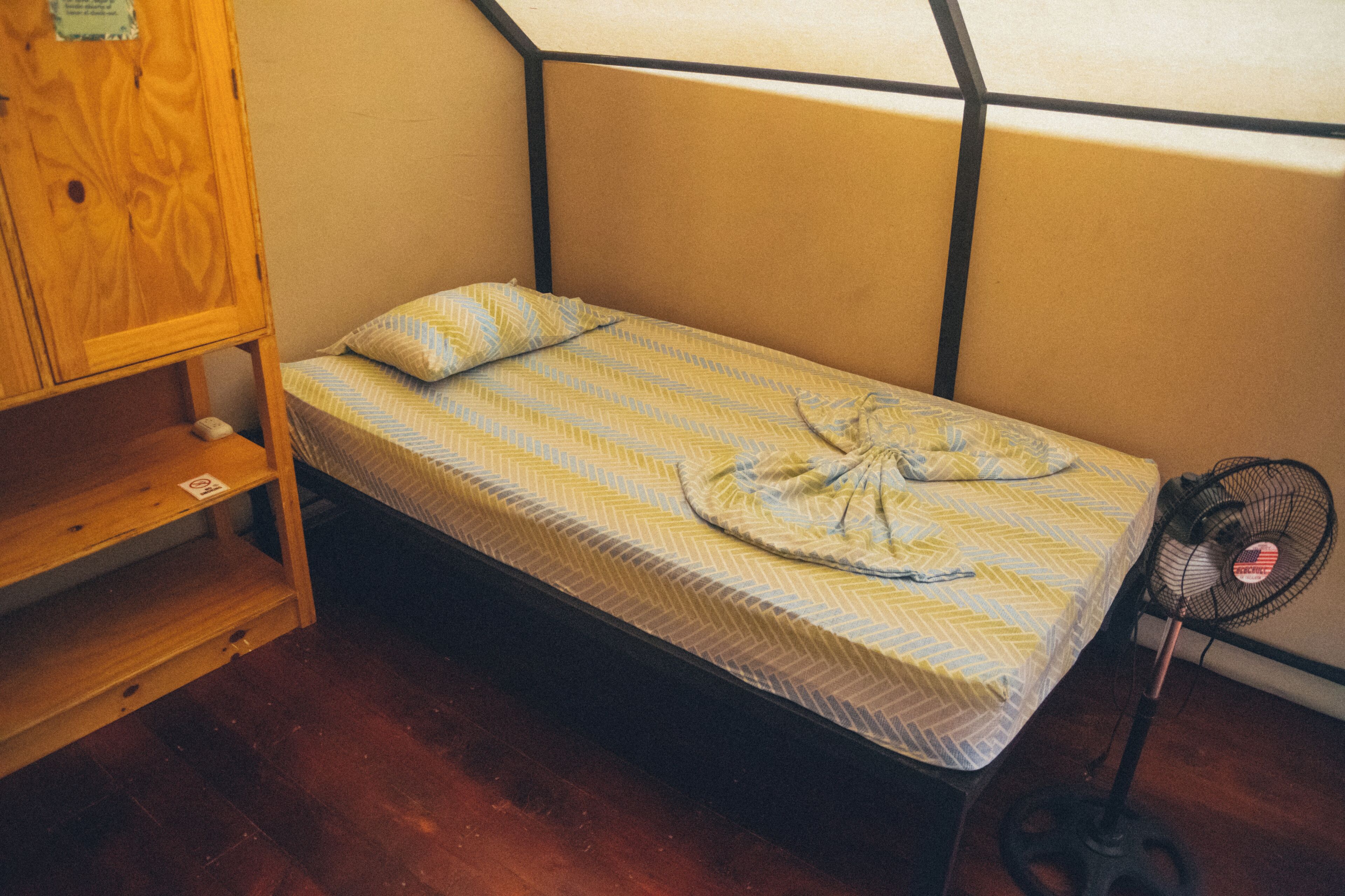Un lit simple avec un matelas à motifs, une armoire en bois et un ventilateur sur pied.