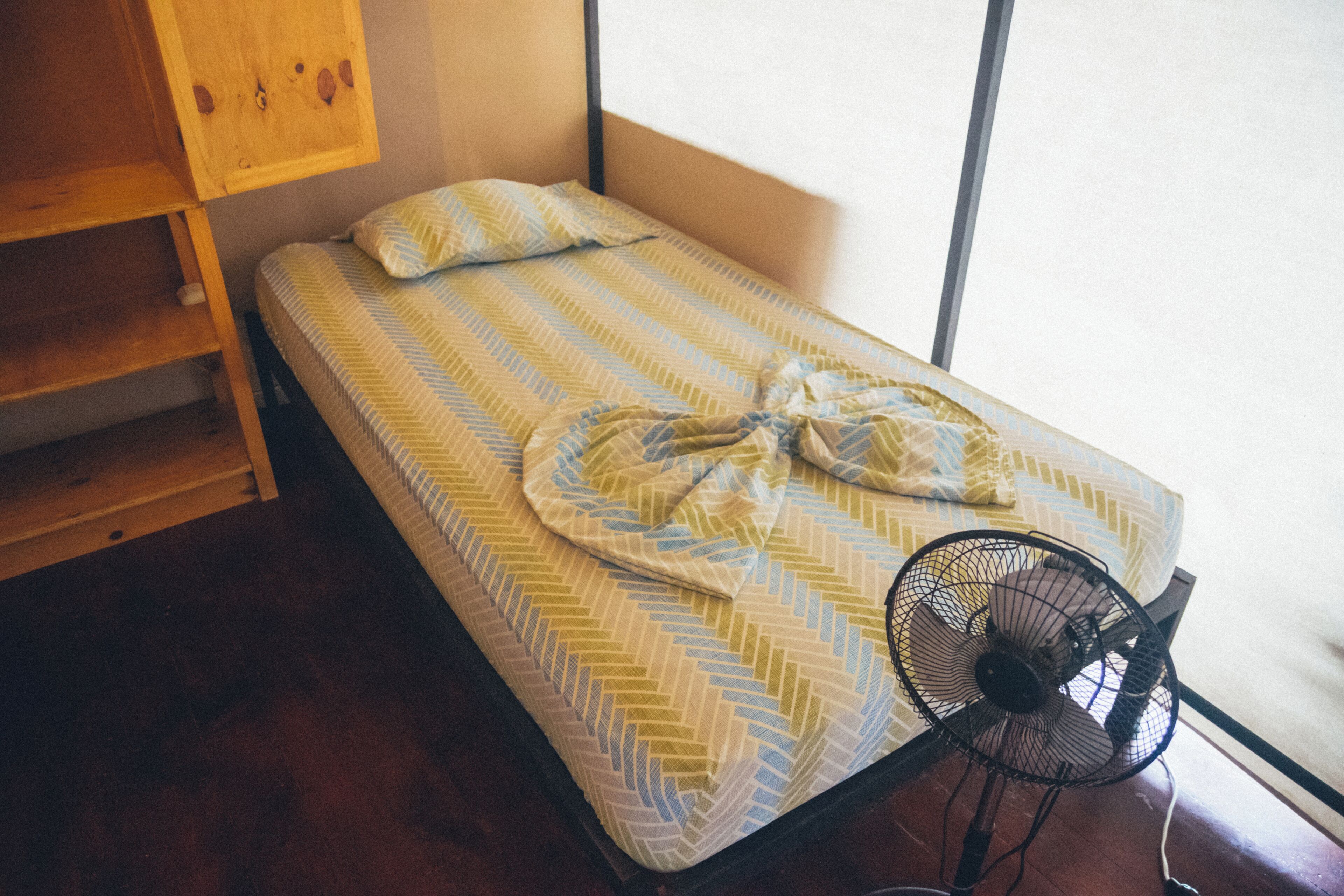 A single bed with a striped bedspread and a fan on the floor.