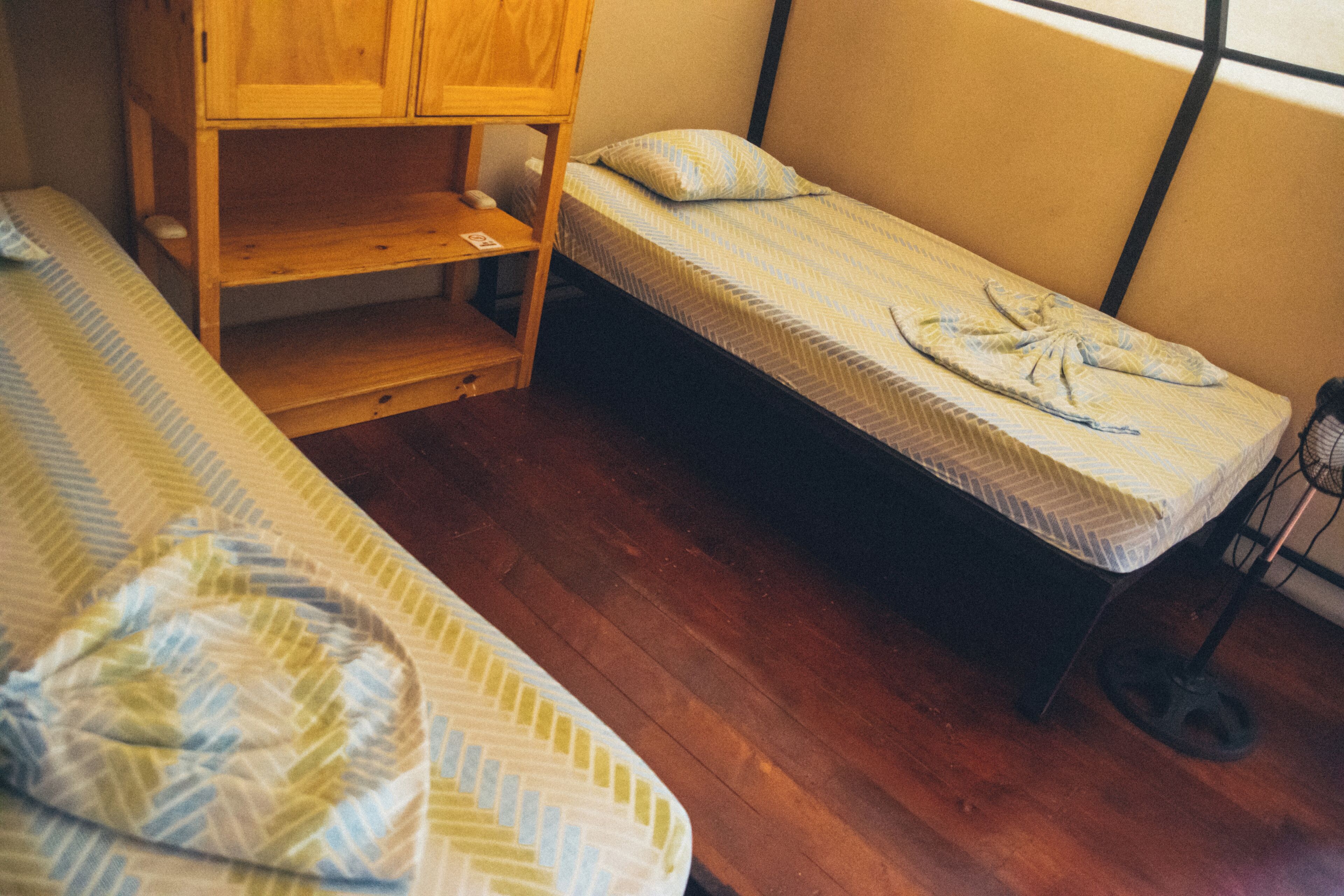 A small, compact room with a single bed, a wooden shelf unit, and a window.