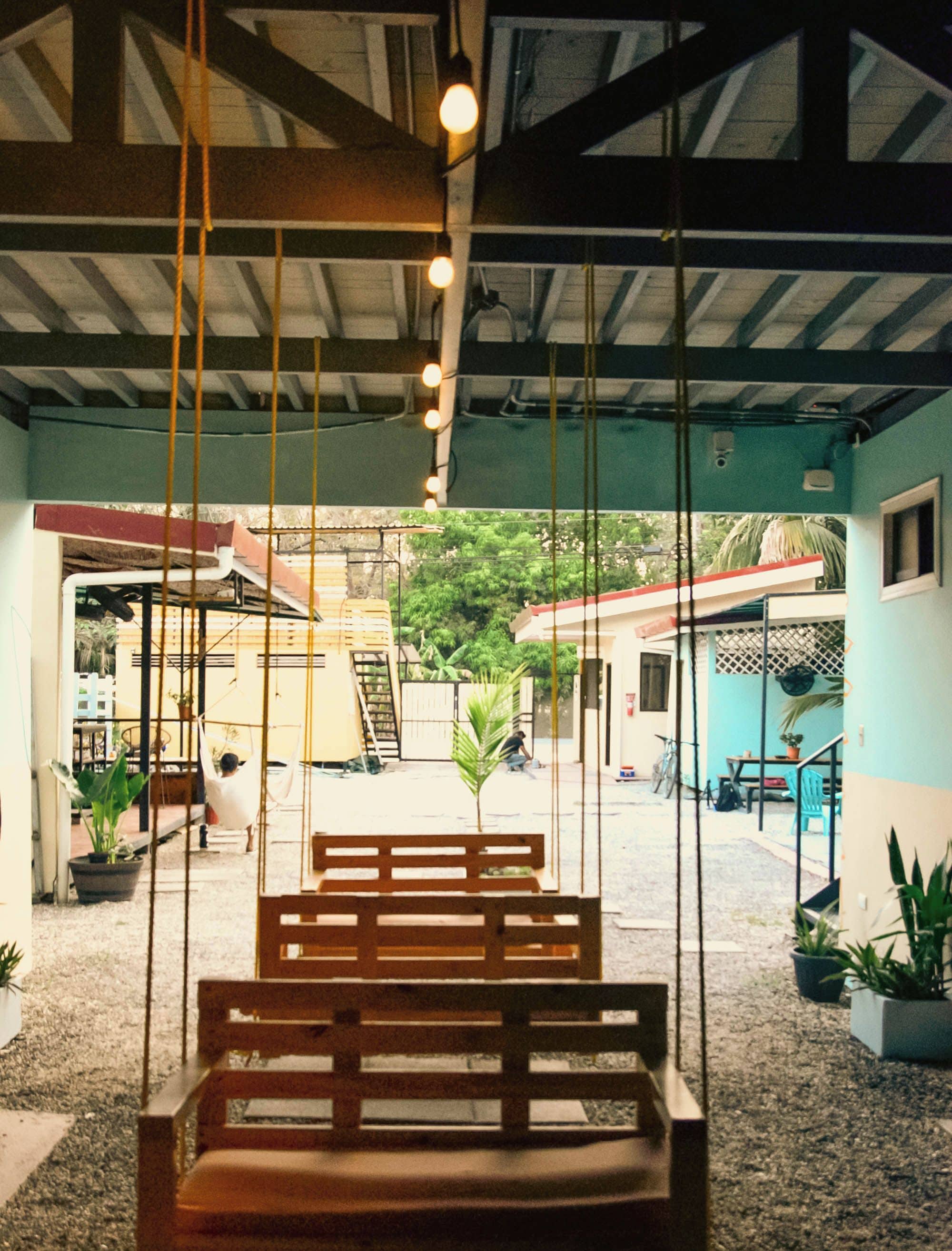 A wooden bench in a covered outdoor area with hanging lights, potted plants, and a view of a building with a red roof.