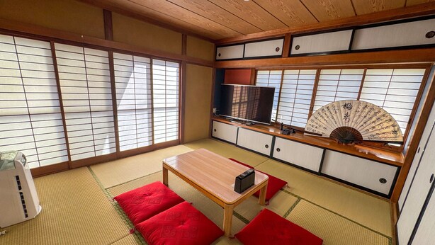 Ett traditionellt japanskt rum med tatamimattor på golvet, ett lågt träbord, röda kuddar, en tv och en dekorativ fläkt.