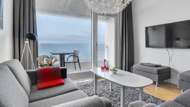 Een moderne woonkamer met een groot raam met uitzicht op zee, een flatscreen televisie aan de wand en een elegante kroonluchter.