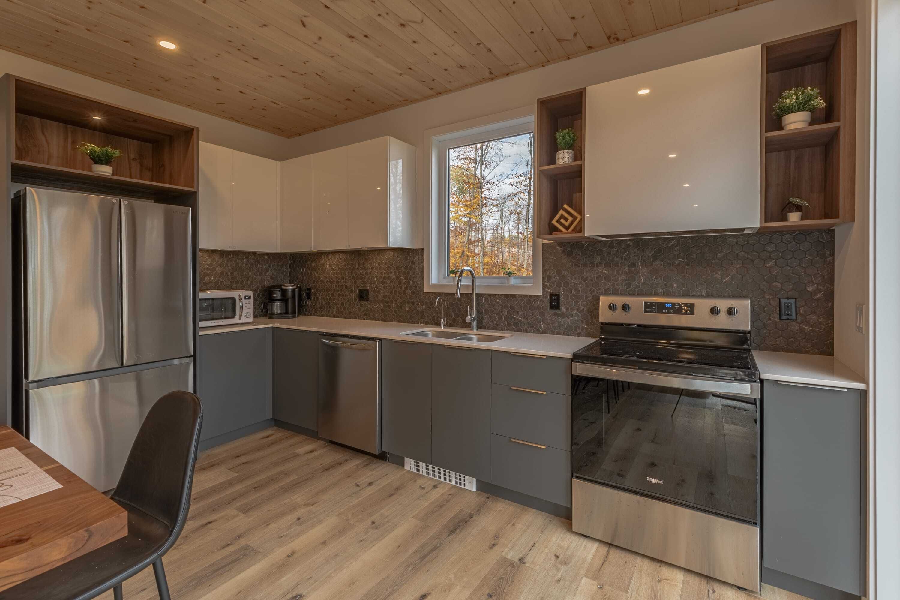 Una cocina moderna con un refrigerador de acero inoxidable, un microondas, un fregadero y un horno. Hay una ventana sobre el fregadero, y estantes de madera con plantas.