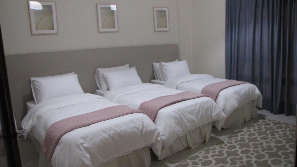 A hotel room with three single beds, each with white bedding and a pink throw blanket. The room has a neutral color scheme with a patterned rug on the floor.