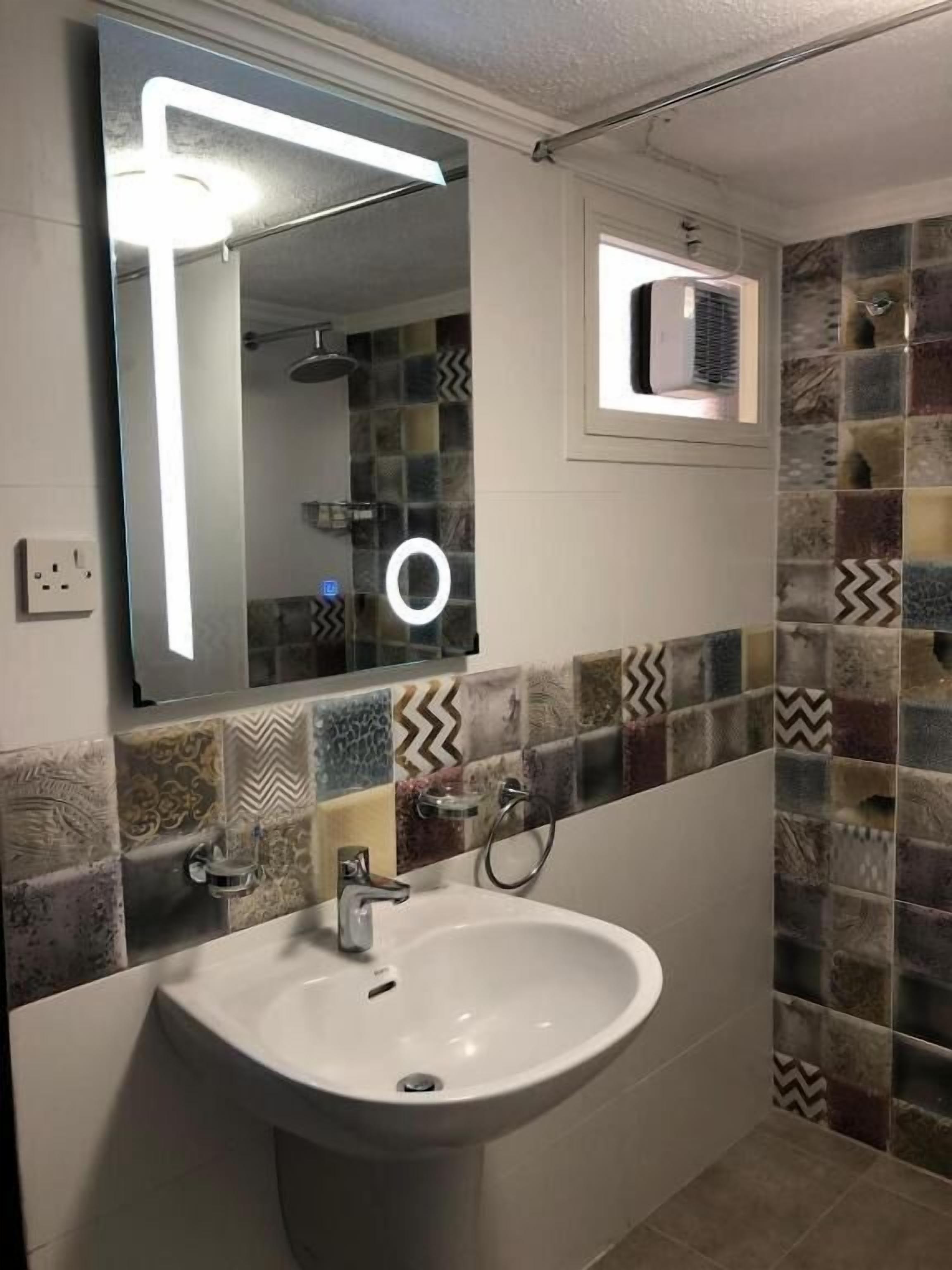 A bathroom with a white sink, a mirror with a light, and patterned tiles.