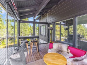 Une terrasse extérieure moderne avec une table en bois, deux tabourets de bar, un canapé rouge et une véranda en verre offrant une vue sur les arbres.