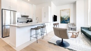 Cocina moderna con armarios blancos, un refrigerador de acero inoxidable y una isla central con taburetes de bar. Hay un televisor de pantalla plana montado en la pared y un cuadro abstracto grande sobre la isla.