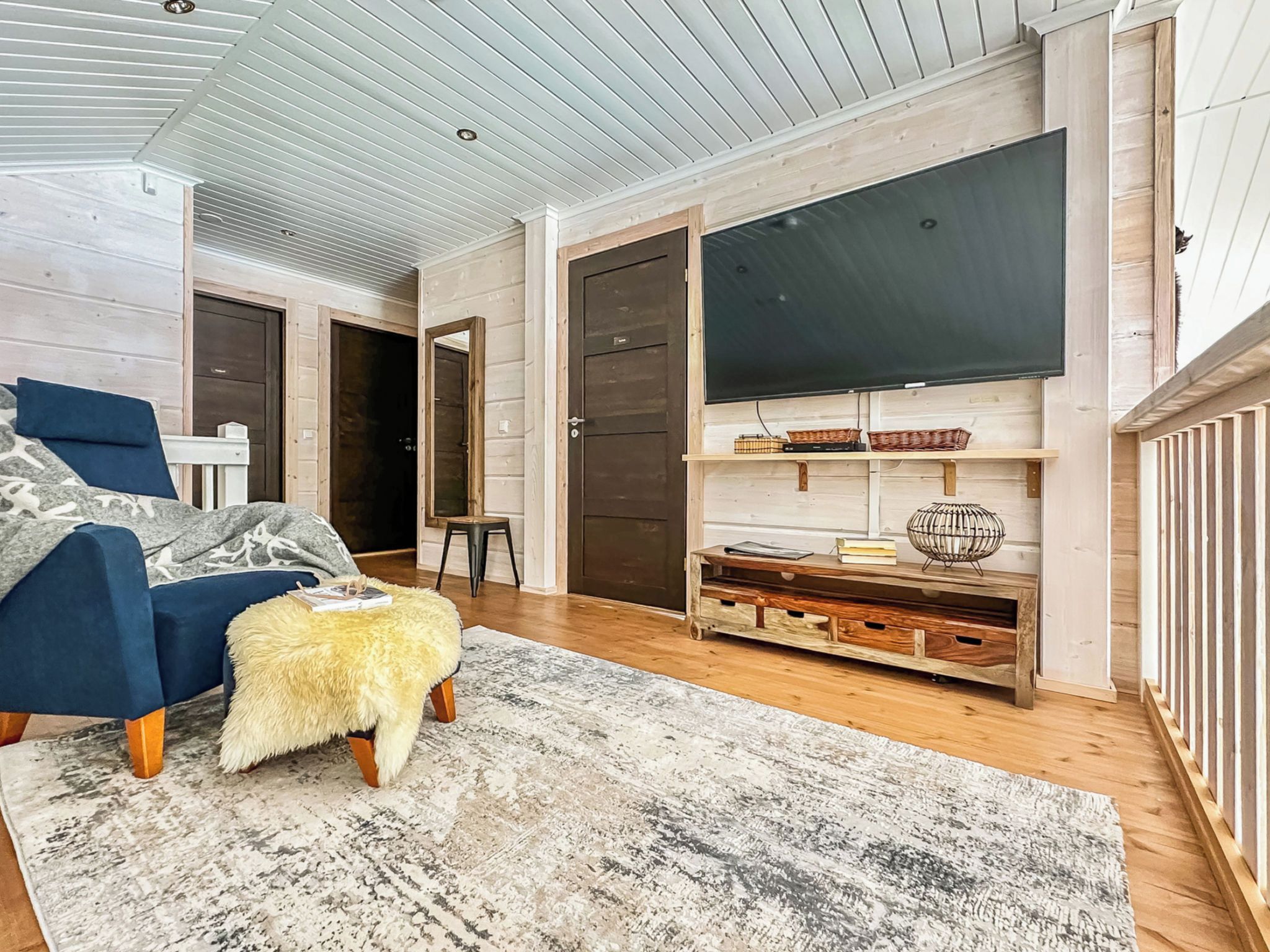 Une pièce avec un fauteuil bleu, un pouf en fourrure, un plancher en bois, un grand téléviseur à écran plat fixé au mur et un meuble rustique en bois agrémenté de paniers.