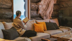 Et hyggeligt interiør i en træhytte med en person, der læser en bog på en sofa, et træbord og et stort vindue, der viser et sneklædt landskab.