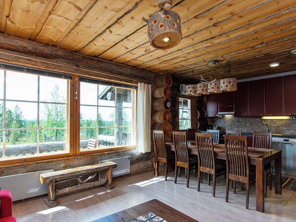 A rustic cabin interior with a dining area, wooden ceiling, large windows, and a stone fireplace.