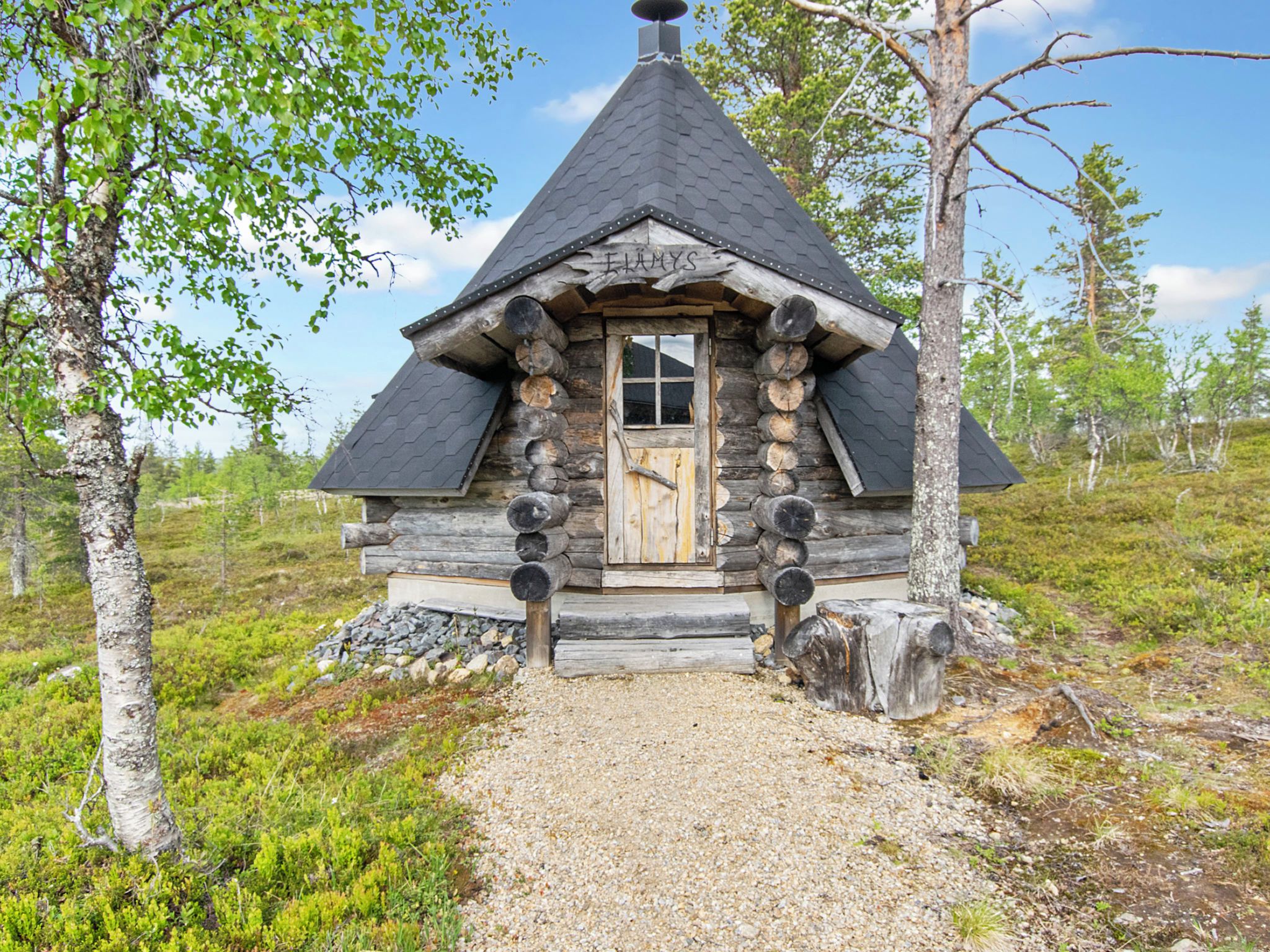 Une cabane en rondins surmontée d’un toit conique, avec une porte en bois et un chemin de gravier menant à l’entrée.