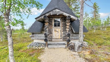 Une cabane en rondins surmontée d’un toit conique, avec une porte en bois et un chemin de gravier menant à l’entrée.