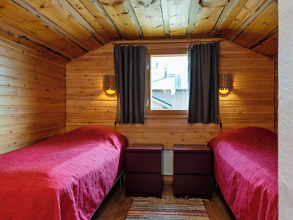 A small, cozy room with wooden walls and a sloped ceiling. There are two beds with red bedding, a small table, and two wall-mounted lamps.