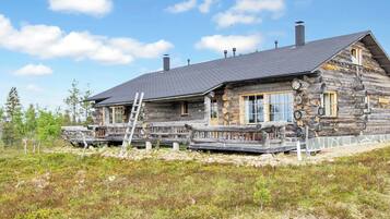 A log cabin with a wooden deck and a ladder, surrounded by natural vegetation.