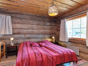 A log cabin bedroom with a bed, two bedside lamps, a window with curtains, and a rustic wooden ceiling.