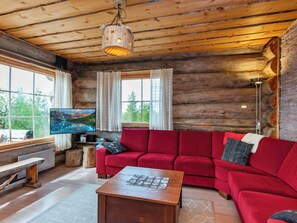 A log cabin interior with a red sofa, a wooden coffee table, a television, and a large window with curtains.