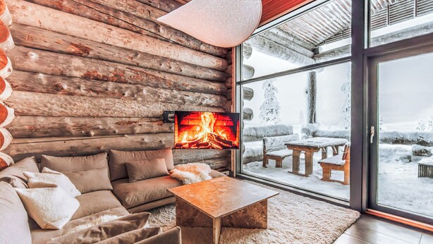 Ein gemütliches Blockhaus-Interieur mit Kamin, bequemen Sitzmöglichkeiten und einem großen Fenster mit Blick auf eine verschneite Landschaft.