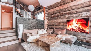 Ein Blockhaushaus-Interieur mit Steinwand, einer Holztreppe, einer Sofagarnitur, einem weißen, flauschigen Teppich und einem Kamin mit Fernseher.