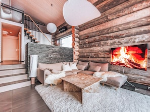 A log cabin interior with a stone wall, a wooden staircase, a sofa set, a white furry rug, and a fireplace with a TV.