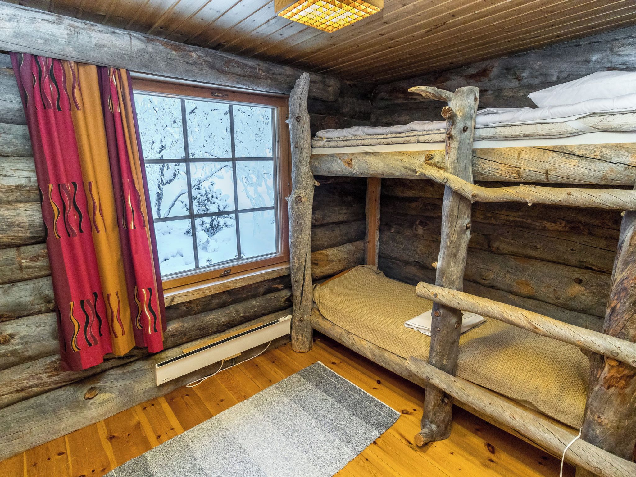 L’intérieur d’une cabane avec un lit superposé, une fenêtre donnant sur des arbres couverts de neige et un radiateur posé sur le sol.