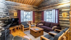 A log cabin interior with a stone fireplace, wooden furniture, and large windows.