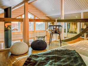 Ein Zimmer mit hohen Decken, großen Fenstern, einem Bett, einem Sessel mit Stofftieren und einem gestrickten Hocker.