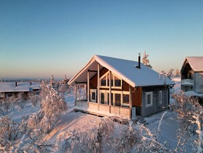 Une cabane en bois, dotée d’un toit en pente et de grandes fenêtres, entourée d’arbres couverts de neige.