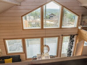 A cabin with a loft bedroom featuring large windows, a dining area with a table and chairs, and a view of the surrounding forest.