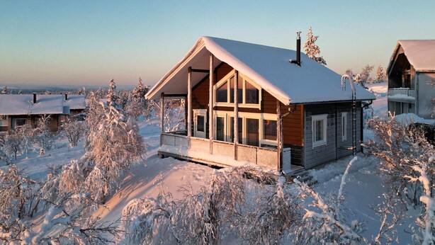 A wooden cabin with a large porch and multiple windows, surrounded by snow-covered trees and a clear sky.