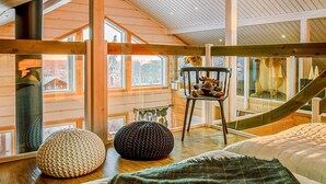 A cozy cabin interior with a loft bed, a chair with stuffed toys, and a view of the outside through large windows.