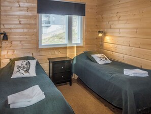 A room with two beds, a nightstand, a window with blinds, and a wall-mounted lamp.