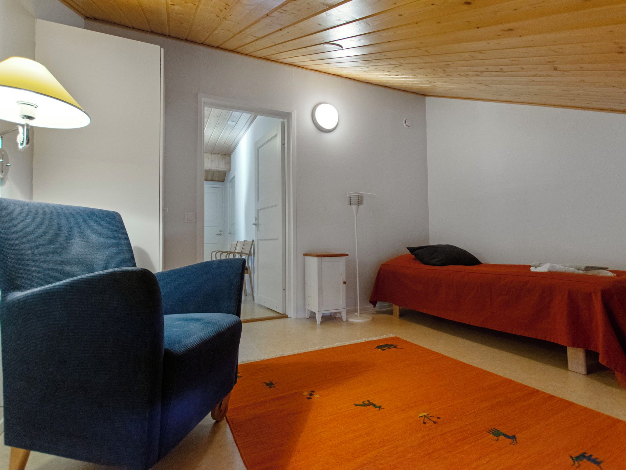 Une pièce avec une chaise bleue, un lit recouvert d’une couverture rouge, une petite table blanche et un lampadaire.