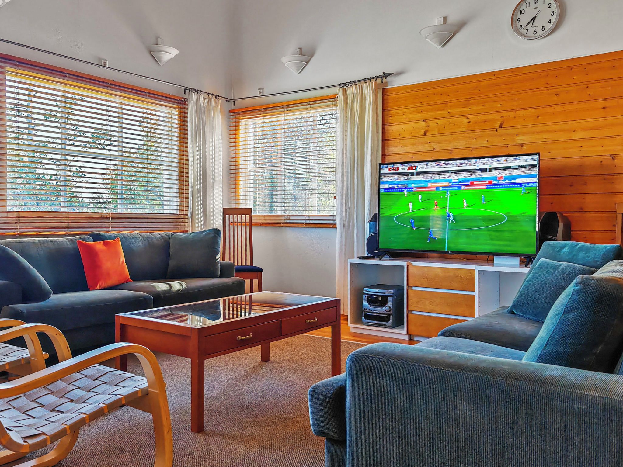 A living room with a sofa, armchair, coffee table, TV, and windows with blinds.