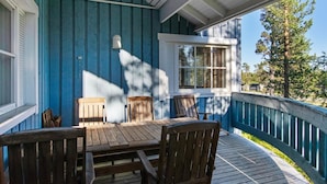 Une véranda couverte avec une table et des chaises en bois, une fenêtre avec des volets, et une vue sur des arbres et une maison au loin.