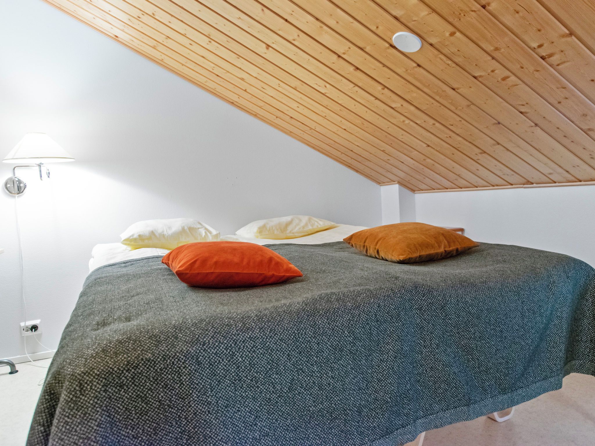 Een slaapkamer met een schuine kap, een bed met een grijze deken en twee kussens, een oranje en een bruin.
