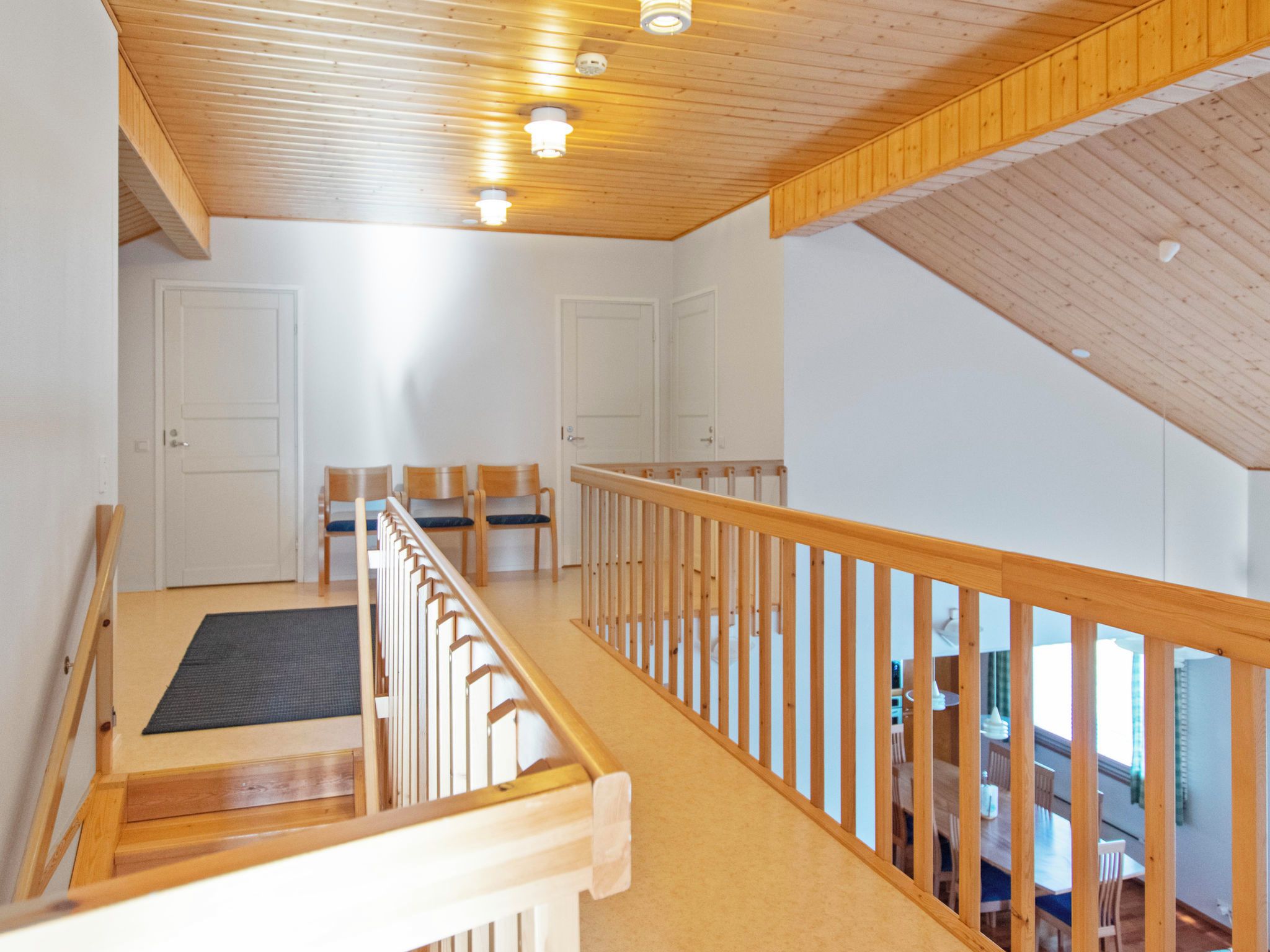 A spacious room with a wooden ceiling, a balcony with a railing, and a view of a dining area.
