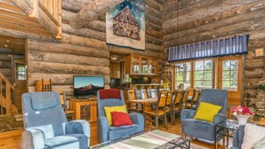 Interior de una cabaña de madera con comedor, dos sillones, un televisor y una cocina visible a través de una puerta abierta.