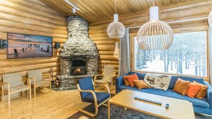 A cozy log cabin interior with a stone fireplace, wooden furniture, and a large window offering a view of a snowy forest.