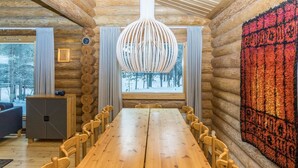 A log cabin interior with a large wooden dining table, chairs, a hanging light fixture, and a view of a snowy forest through the windows.