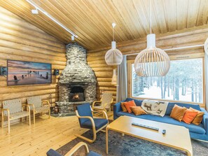 A log cabin interior with a stone fireplace, wooden furniture, and a large window with a view of trees.
