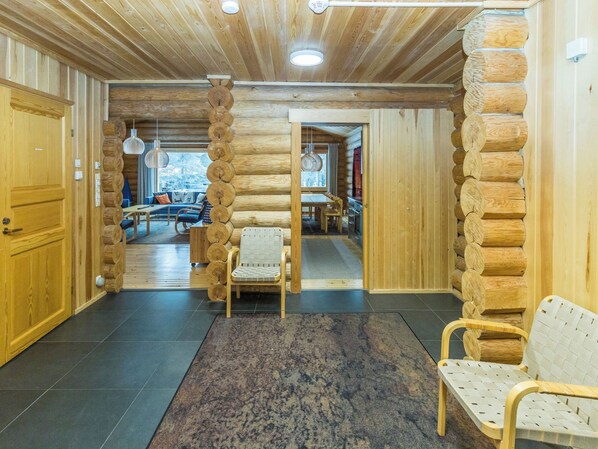 A log cabin interior with a central seating area, wooden walls, and a view to a dining area.