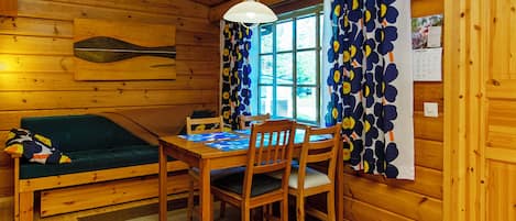 A wooden cabin interior with a dining table, chairs, a sofa, and a window with curtains.