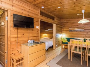 Interior de una cabaña de madera con una cama, un escritorio, un televisor montado en la pared y un comedor con sillas.